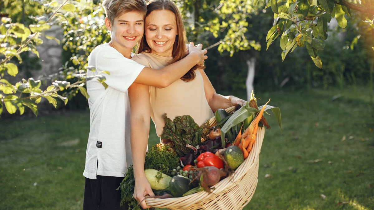 Mother and son smiling outdoors with a basket full of fresh vegetables, embracing in a sunny garden.